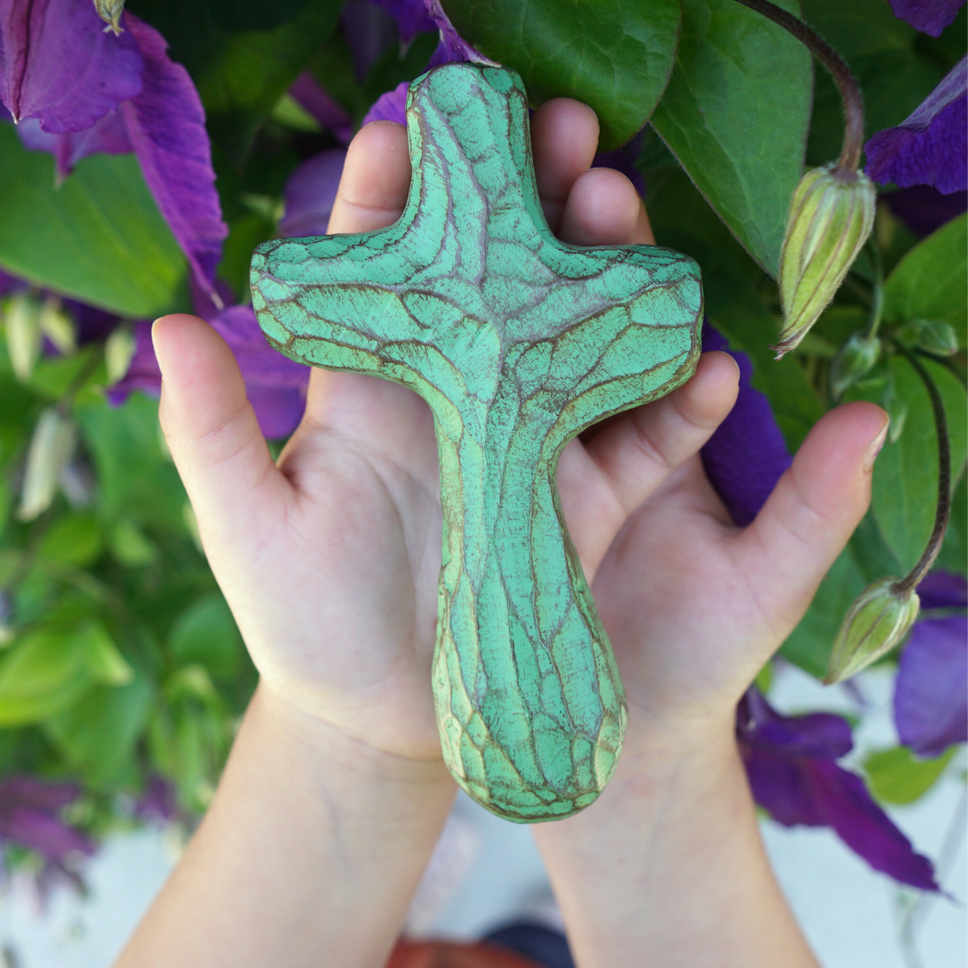 Hands holding patina green painted wooden holding cross in front of purple flowering plant